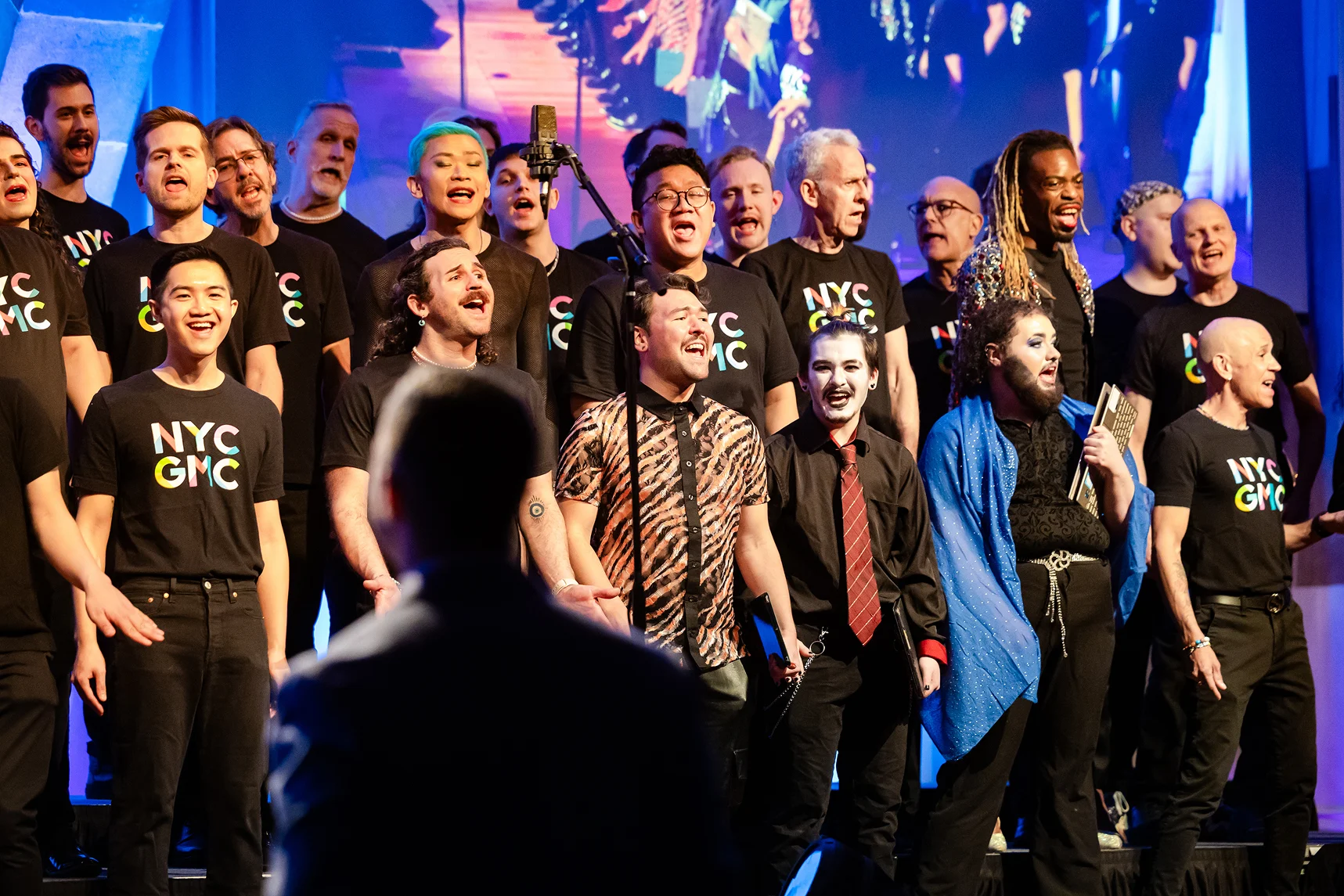 A choir performs on stage under blue stage lighting, singing energetically toward a microphone stand.