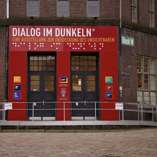 Entrance to "Dialog im Dunkeln" exhibition with a red facade and large windows, located in Block W of a brick building.