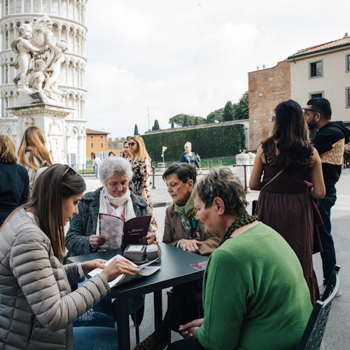 Lidé sedí u venkovního stolu kavárny poblíž Šikmé věže v Pise, zatímco v pozadí ostatní procházejí a komunikují.