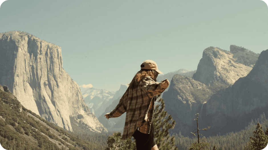 A person wearing a plaid shirt and cap explores the scenic landscape of Yosemite National Park, with iconic granite cliffs and lush green forests visible under a clear blue sky.