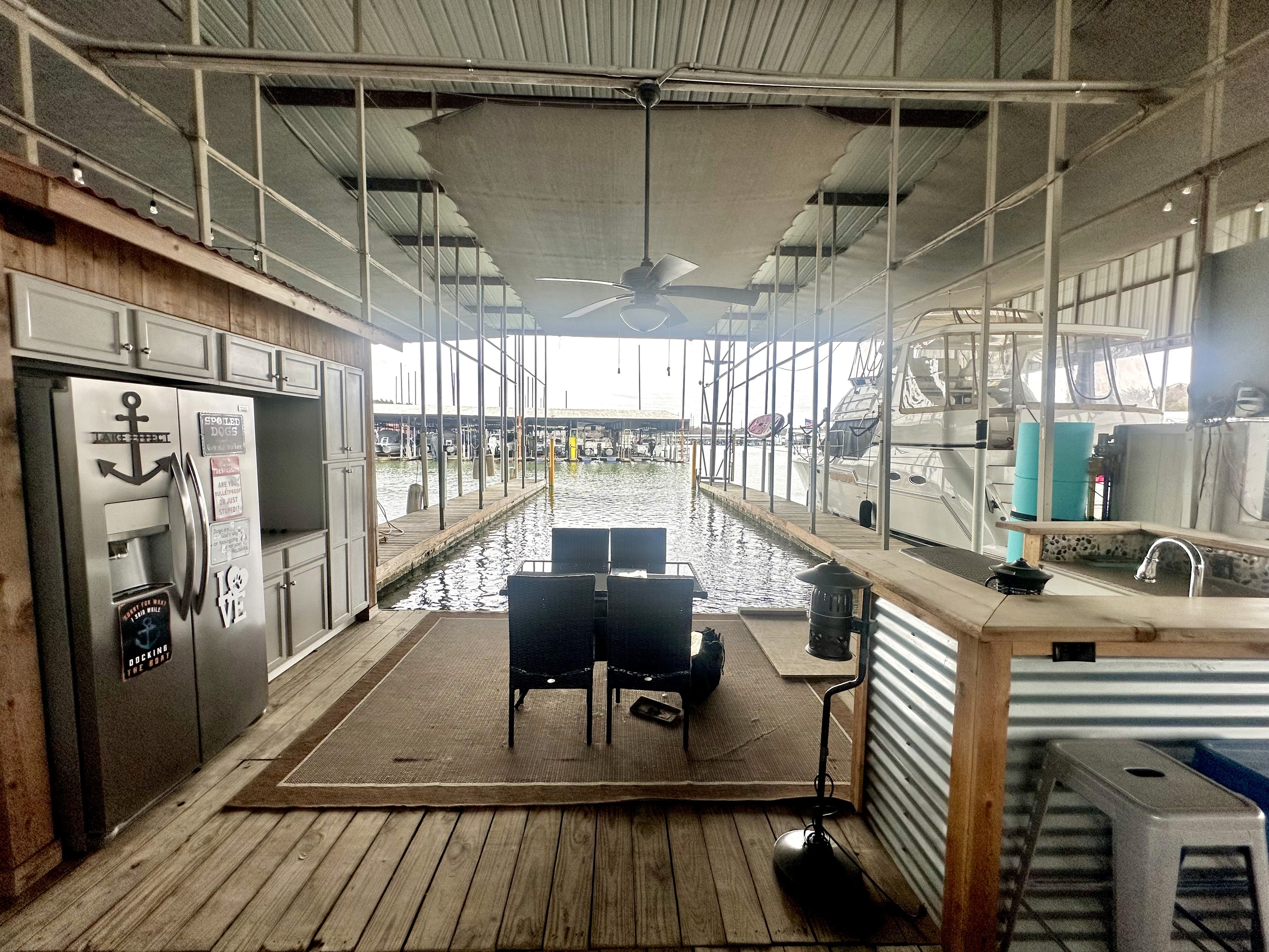 A cozy marina deck features two black lounge chairs facing a serene body of water, with a modern kitchen area on the left, complete with a metallic fridge and nautical decor, all under a spacious, roofed structure.