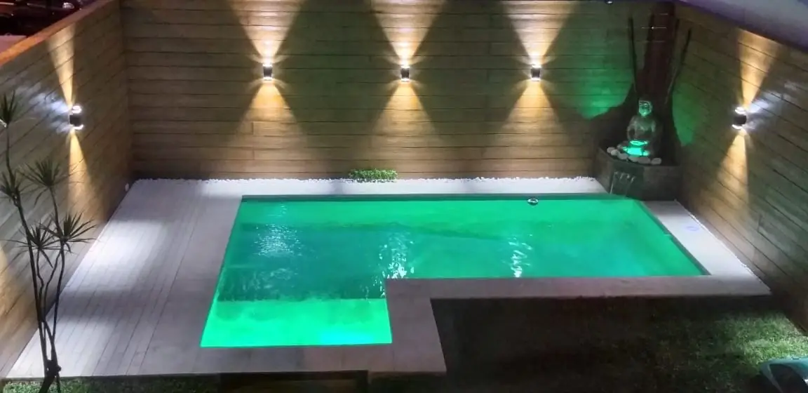 Pileta de diseño en L iluminada con luces LED subacuáticas verdes durante la noche, enmarcada por una pared de madera con modernos apliques luminosos.