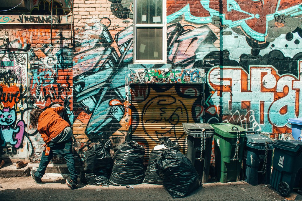 An urban alleyway adorned with vibrant graffiti art on brick walls, featuring a mix of colorful tags and murals. A person in an orange jacket is seen cleaning or organizing black garbage bags near a row of green and blue trash bins. The scene captures the gritty yet artistic essence of city life and street culture.