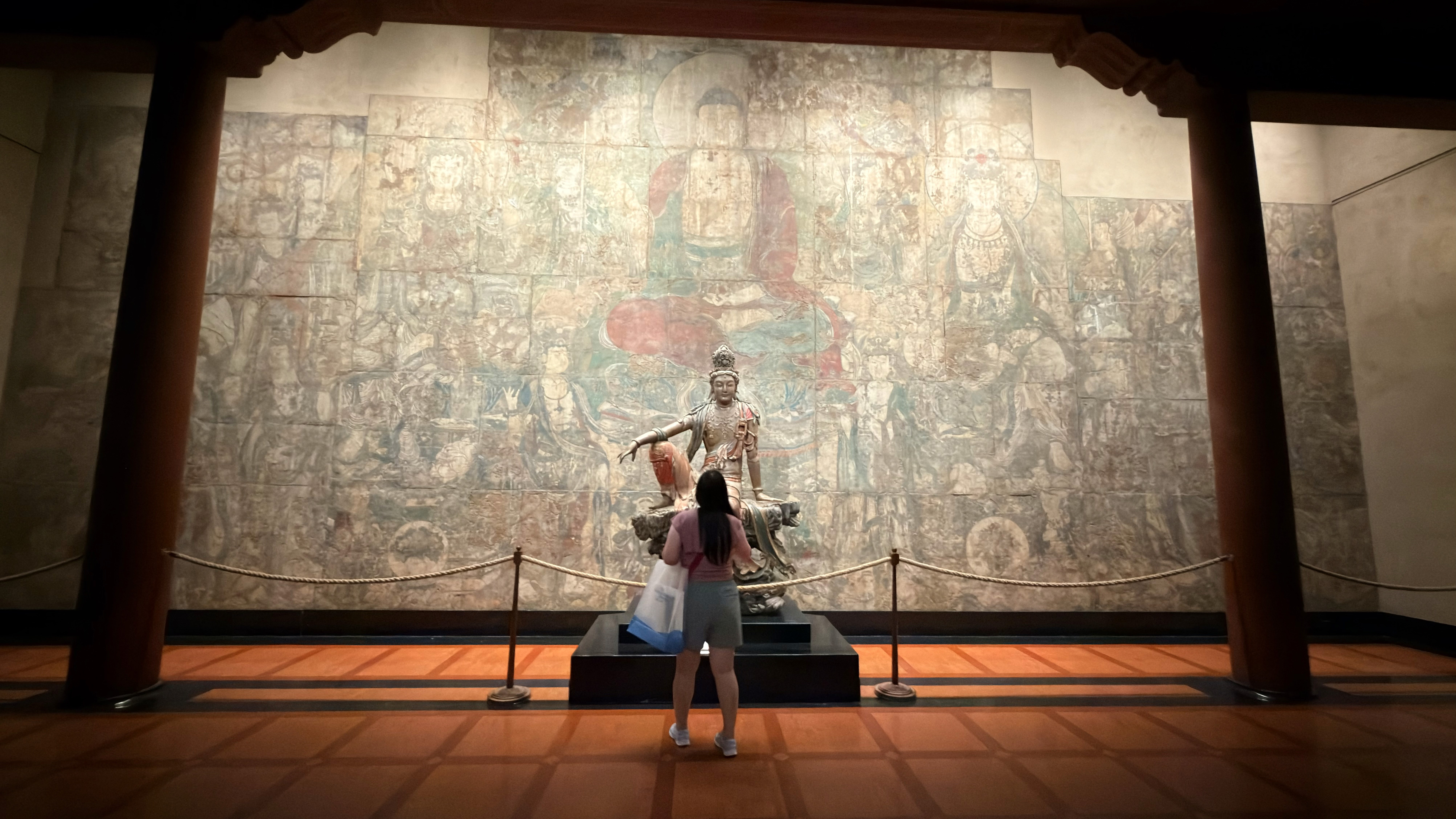 Wide museum-room view of a visitor before Guanyin and a Buddhist mural in the Chinese Temple Gallery