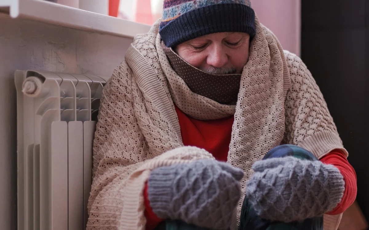A person sits indoors wearing layered winter clothing, including a knitted scarf, jumper, and gloves, beside a radiator. The scene conveys cold weather and the need to keep warm at home.