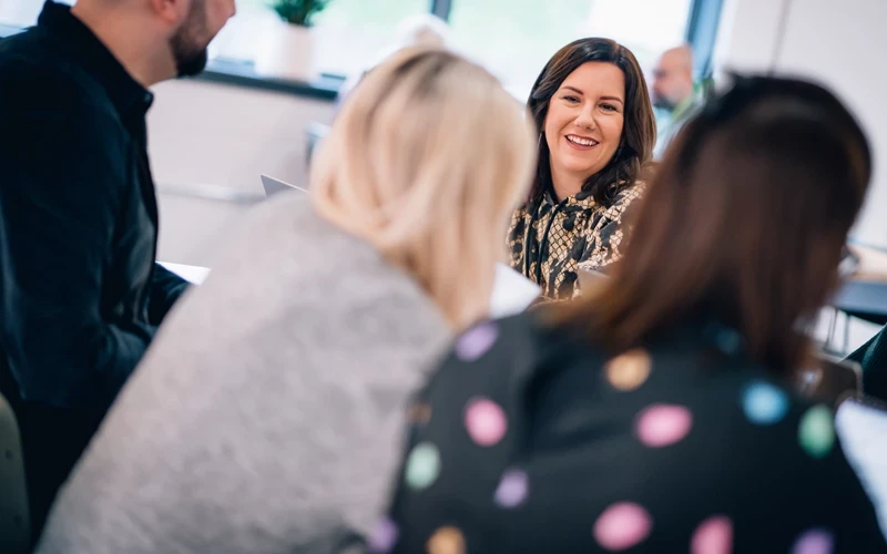 Group of people seated around a table in a bright office space, engaged in discussion with papers in hand.