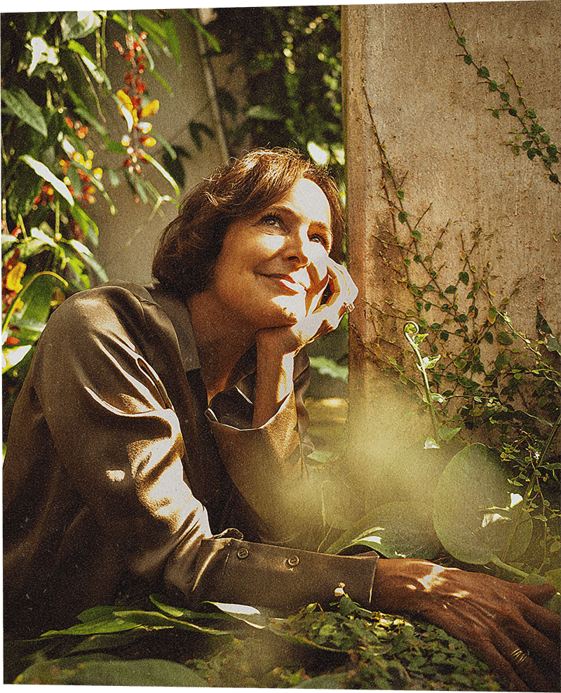 A woman with chin-length brown hair gazes upward in a sunny terrace, leaning her head against her hand.