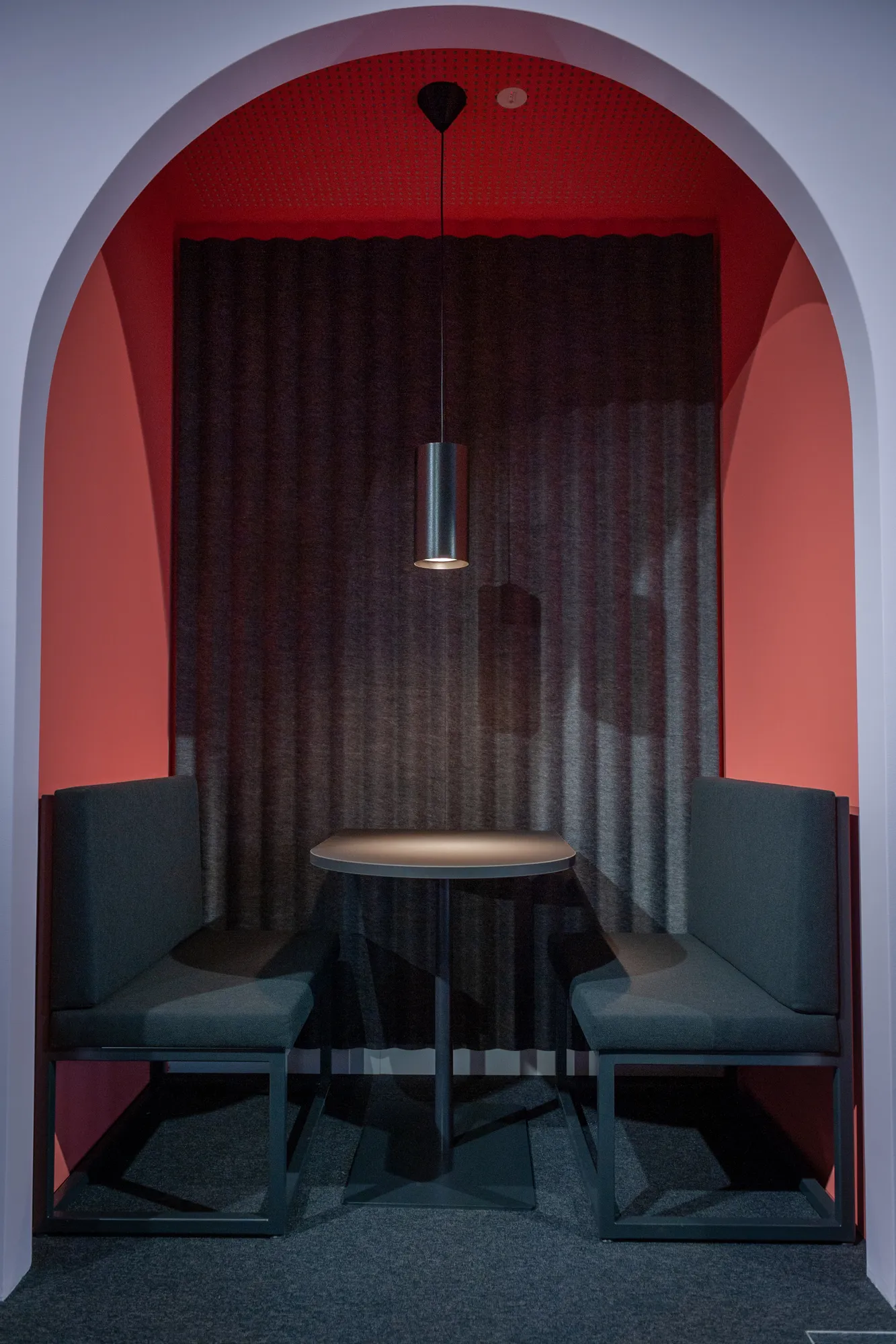 Cosy red arched booth with two grey armchairs, a round table and pendant light.