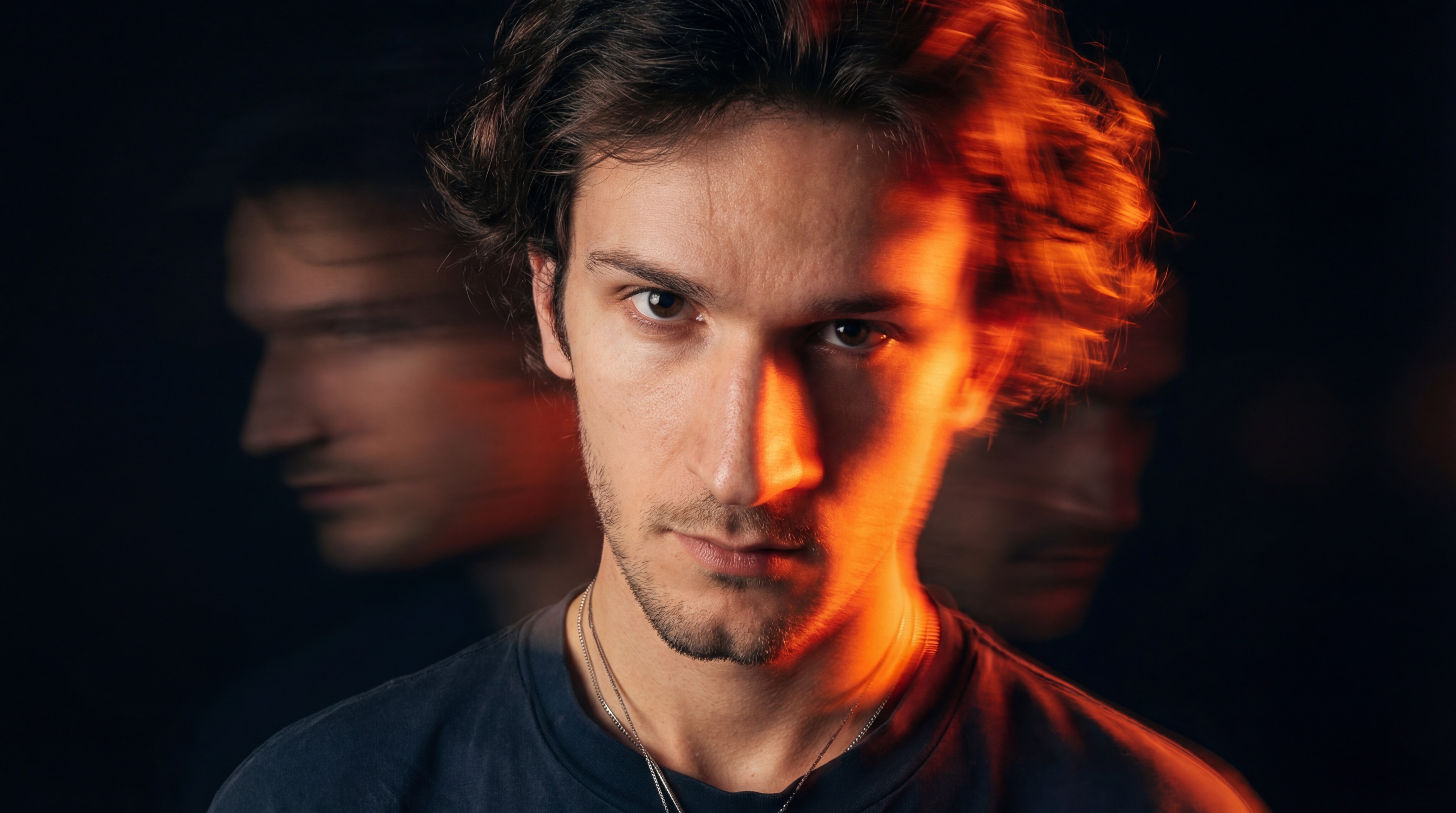 Artistic portrait of young man with curly hair in black shirt and chain, multiple exposure with orange-golden light and neon blue streaks, dynamic motion blur on black background.