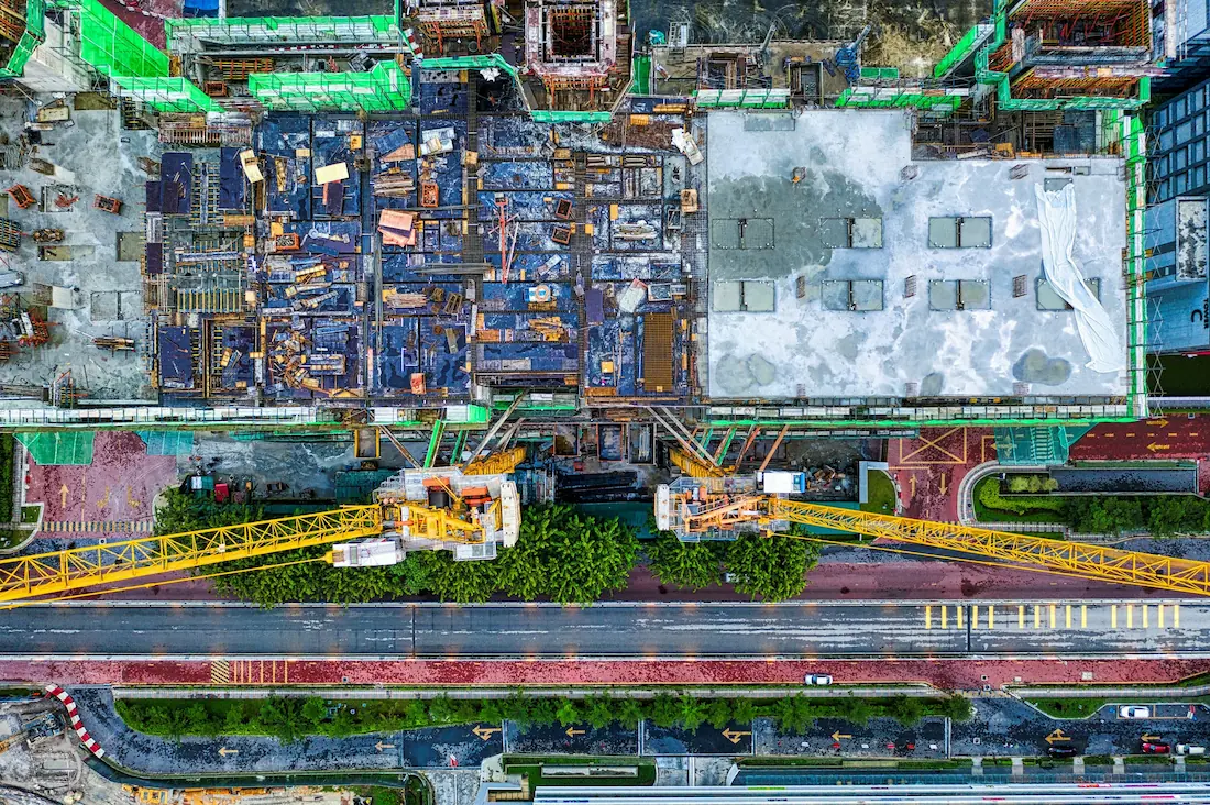 High-angle aerial photography showing a large urban construction layout with yellow cranes over a city road