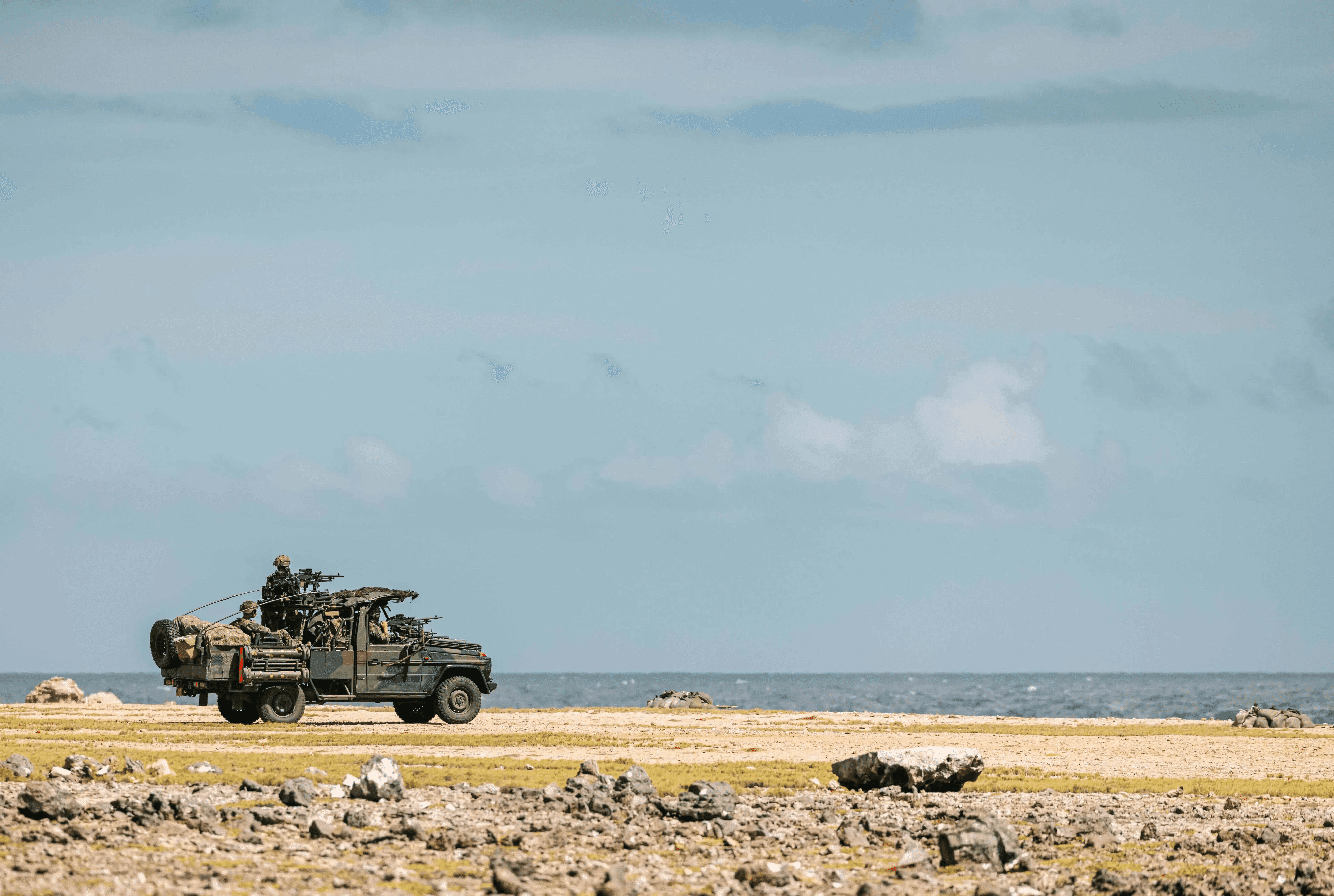 A military vehicle drives along a rocky coastal landscape with a clear blue sky in the background, highlighting an open terrain and ocean view.