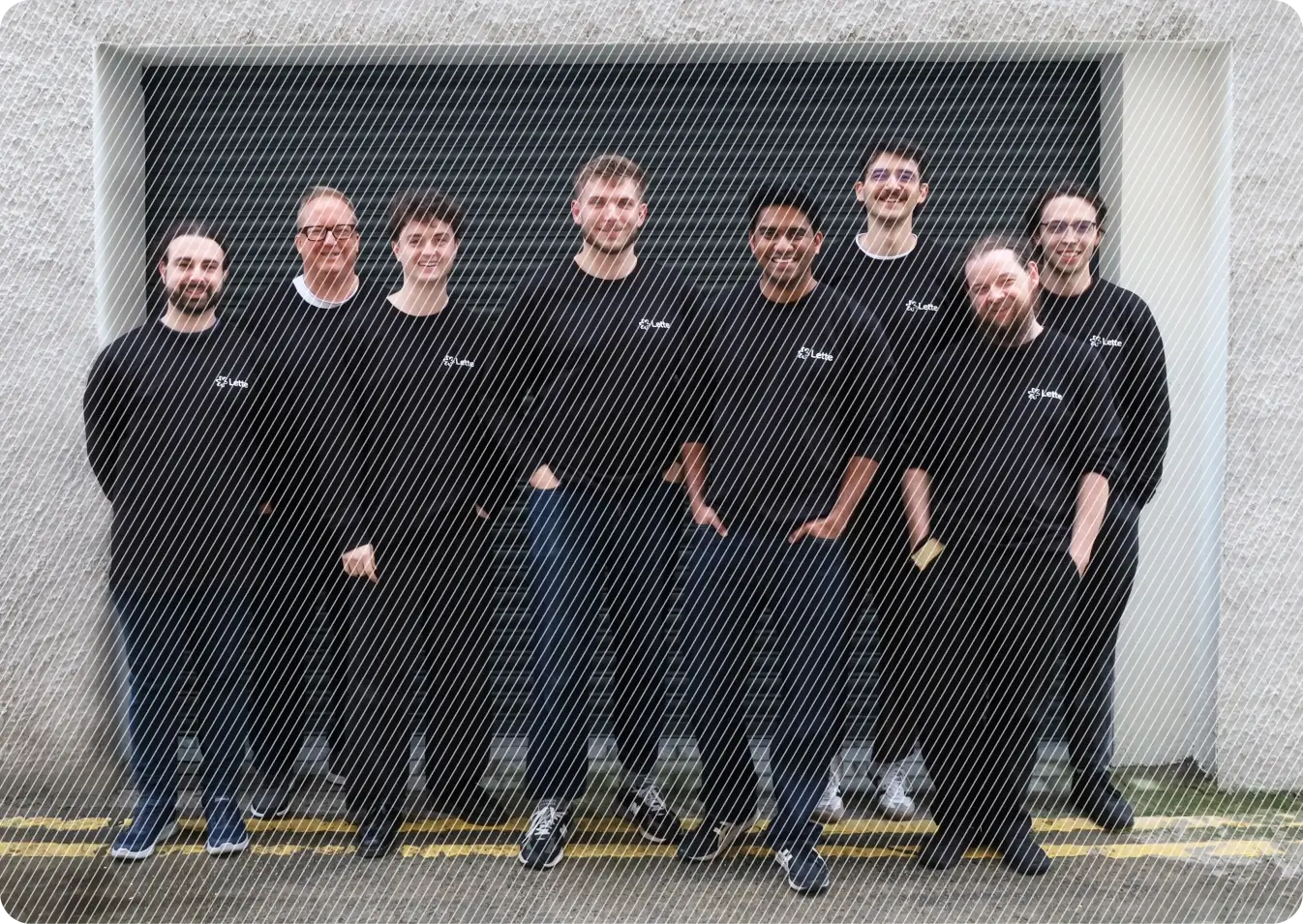 A group of eight people wearing black shirts, standing in front of a large gray garage door, posing for a team photo on a rainy day with a yellow line visible on the wet pavement.
