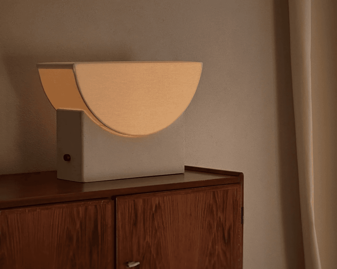 A modern, minimalist lamp with a semi-circular design emits a warm, soft glow, placed atop a wooden cabinet against a neutral-colored wall, creating an elegant and cozy ambiance in the room.