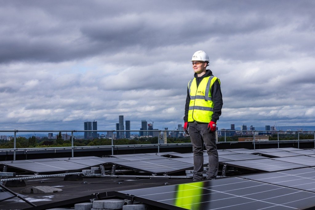 A technician in a high-visibility vest and safety helmet stands proudly on a rooftop covered in solar panels, overlooking a city skyline under a cloudy sky, symbolizing progress in urban renewable energy initiatives.