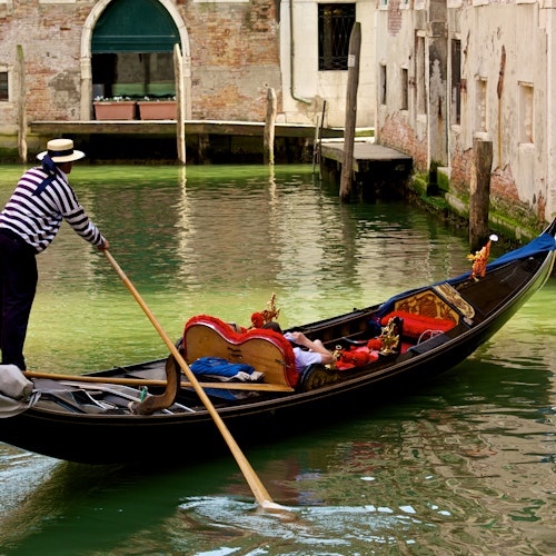 Person in a striped shirt rowing a gondola with cushioned seats along a canal lined with old buildings.