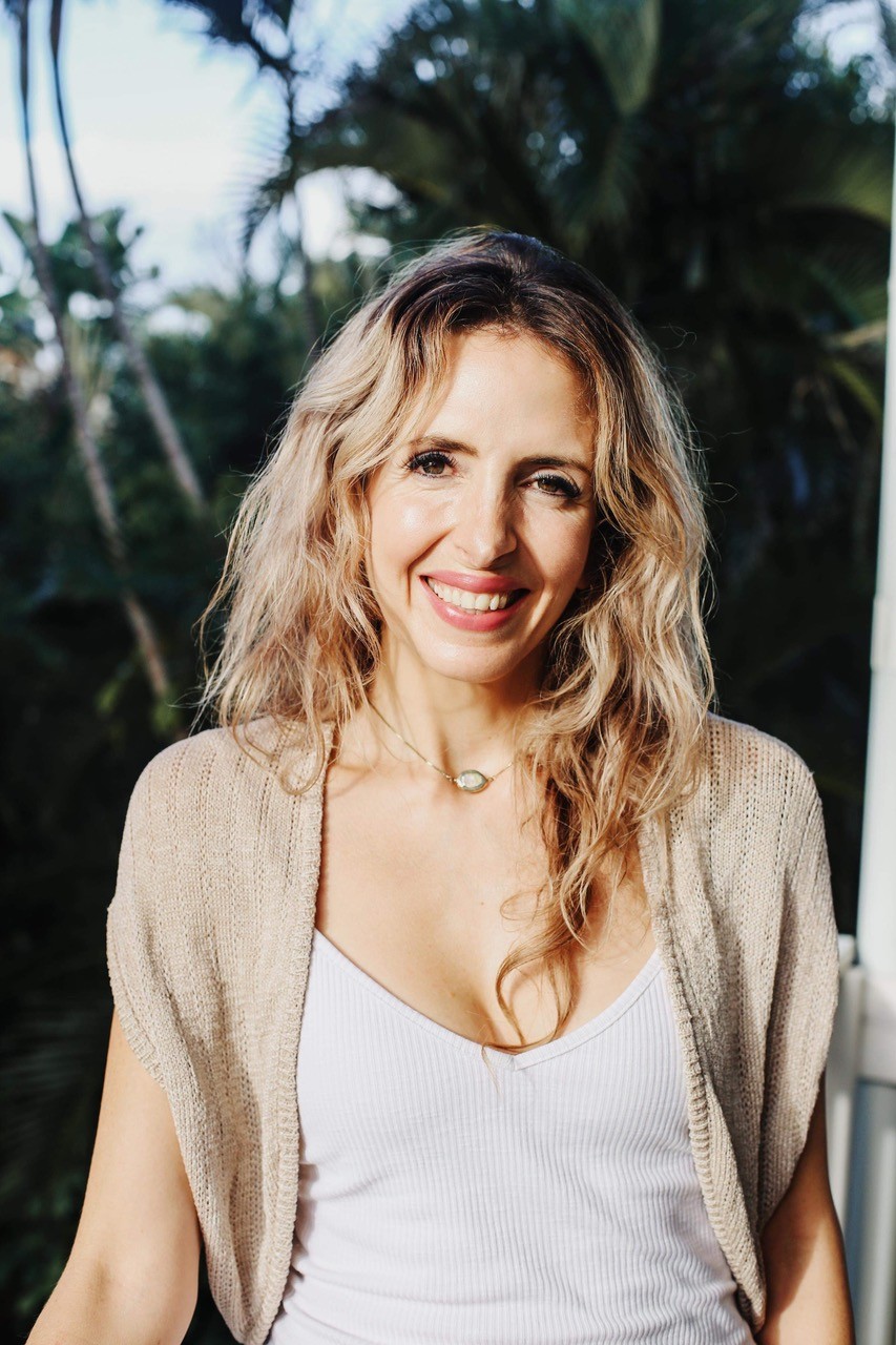 A portrait of Caryn Holzman - smiling woman with wavy blonde hair, wearing a light top, standing outdoors with palm trees in the background.