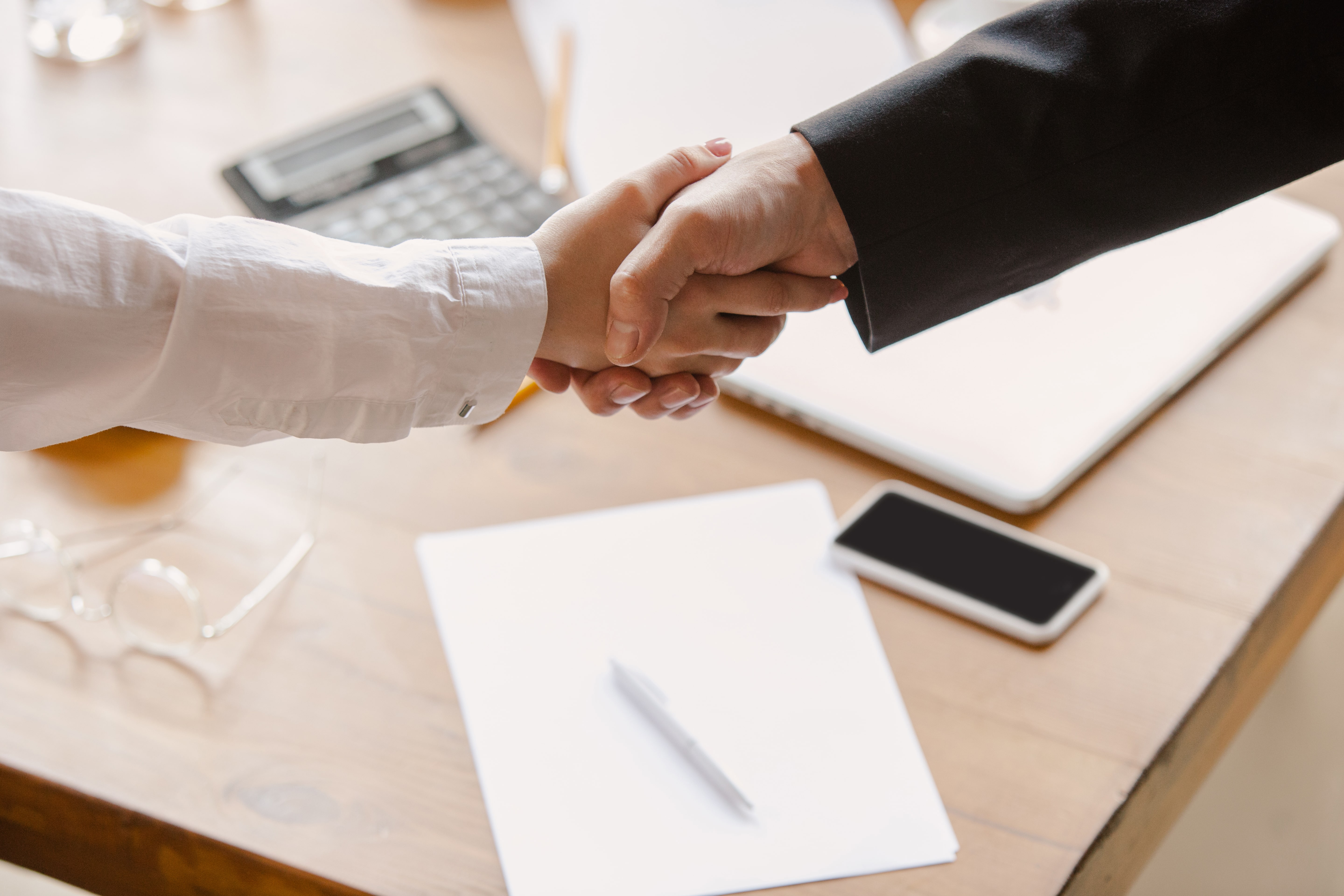 Close-up of a professional business handshake between two people in formal attire at a desk with documents, smartphone, and laptop — symbolizing trust, partnership, and successful business deal.