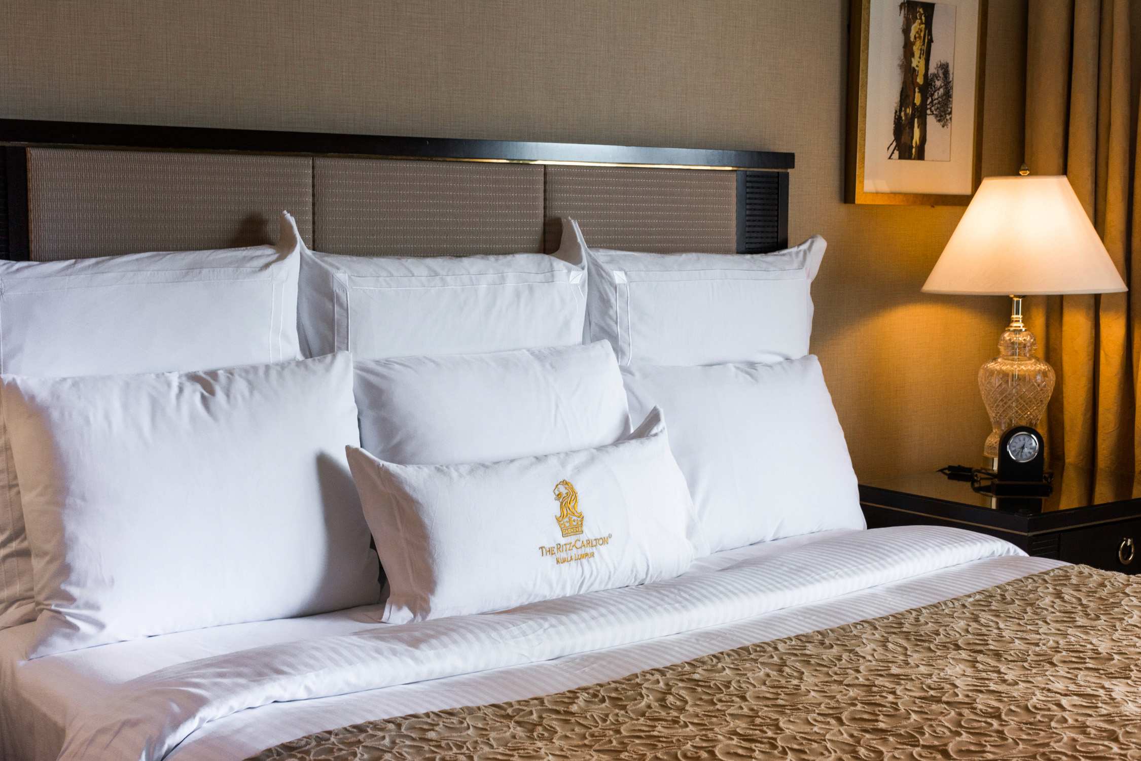 A hotel bed with white pillows and a decorative bolster pillow featuring a gold crest sits against a headboard.