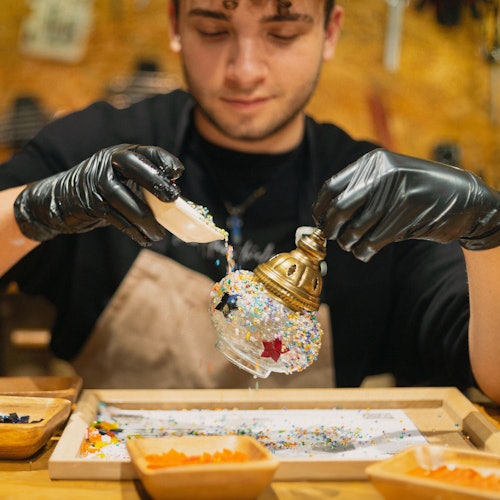 Ang kalahok na may suot na itim na guwantes ay gumagawa ng Turkish mosaic na lampara, nagbubuhos ng makukulay na bato sa isang glass sphere sa isang workshop na set.