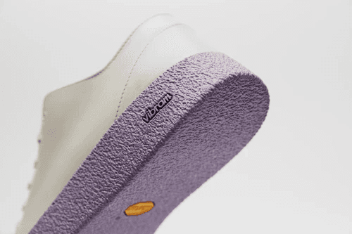 A close-up of a white sneaker showcasing its textured lavender sole, featuring a Vibram logo, highlighting the shoe's stylish and durable design.