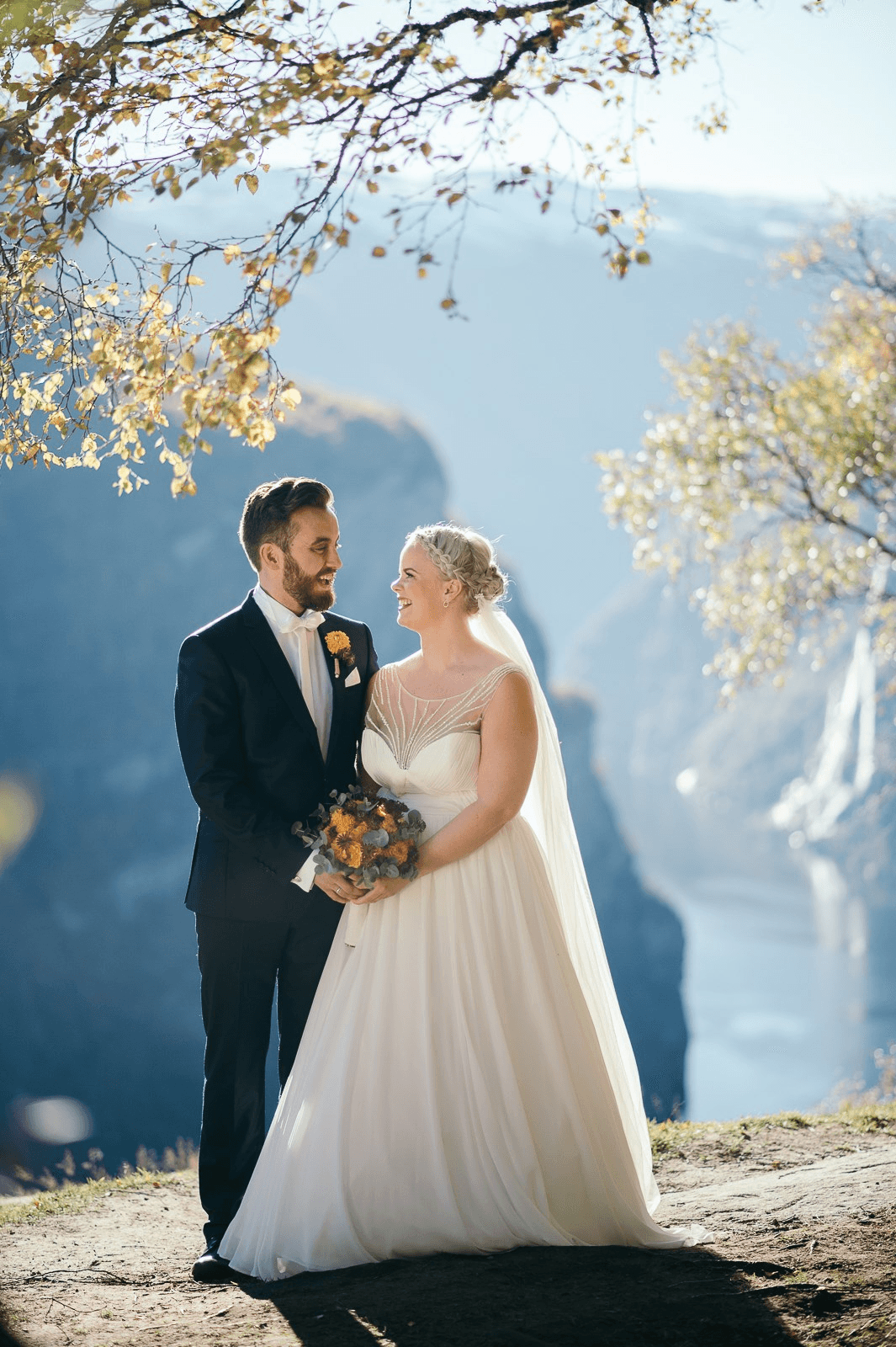 A bride and groom in formal attire stand joyfully under sunlit autumn trees on a cliff edge, overlooking a scenic mountain valley.