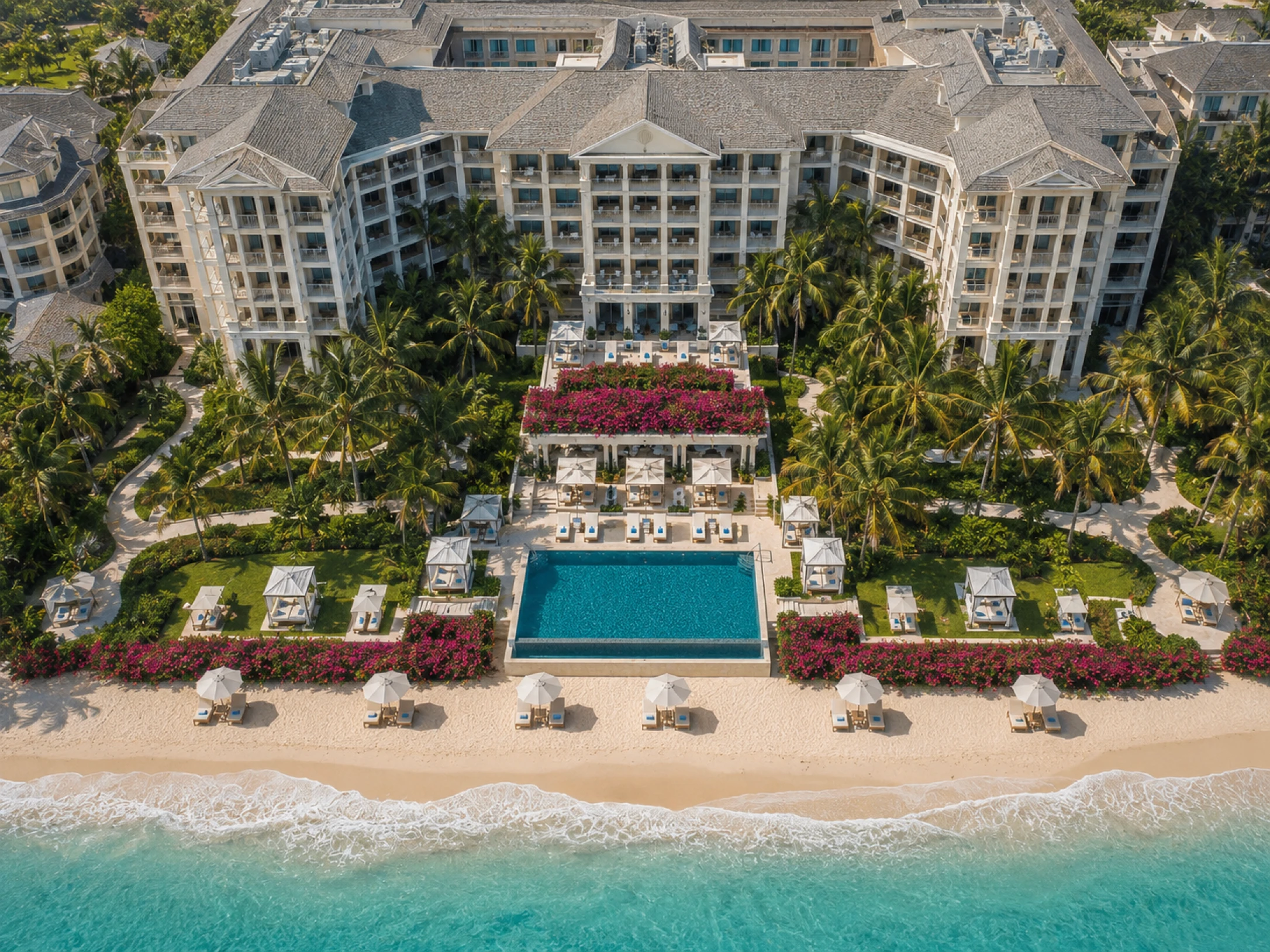 Vista aérea de um hotel de luxo à beira-mar, com piscina, jardins tropicais, áreas de descanso e acesso direto à praia.