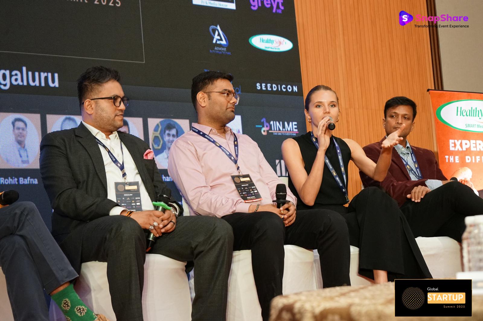 Woman speaking on a panel, holding a microphone and gesturing with her hand, with other panelists seated beside her.