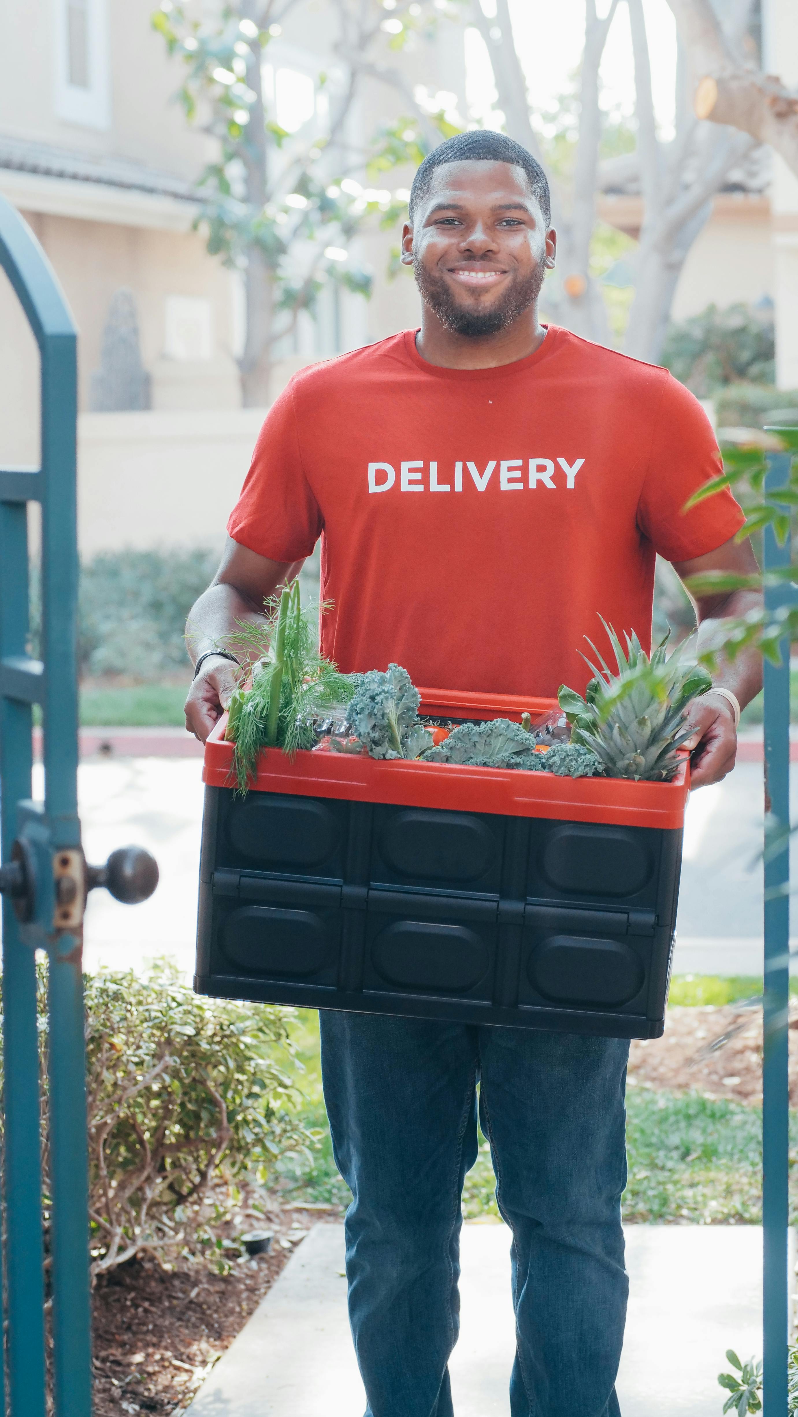friendly-delivery-man-in-a-red-shirt-holding-a-box-of-fresh-vegetables-at-a-fron - kindel-media (pexels)