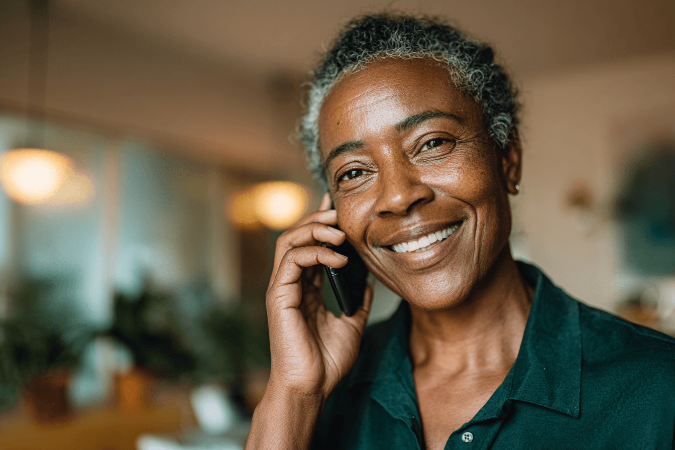a home care agency operator (female, 50s) talking on her cell phone and smiling in a home care agency office. The agency owner is wearing a dark green shirt.