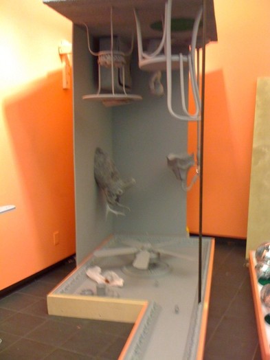 installation of a sculpture made upside-down of a corner of a room with a table, chair, chandelier, a light stand, and a fake deer head mounted on a wall