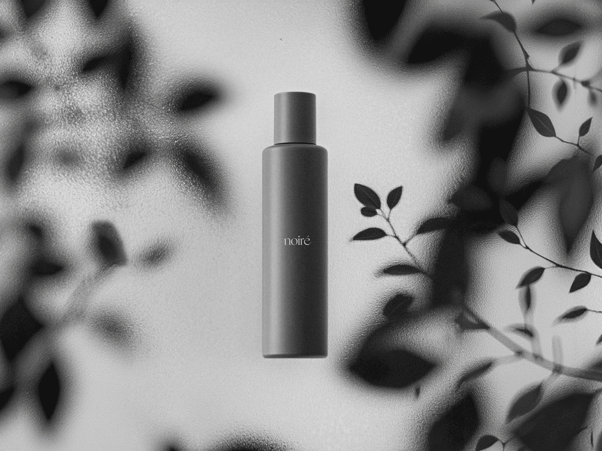 Minimalist black cosmetics bottle with "noirē" branding displayed against frosted glass and leaf shadows