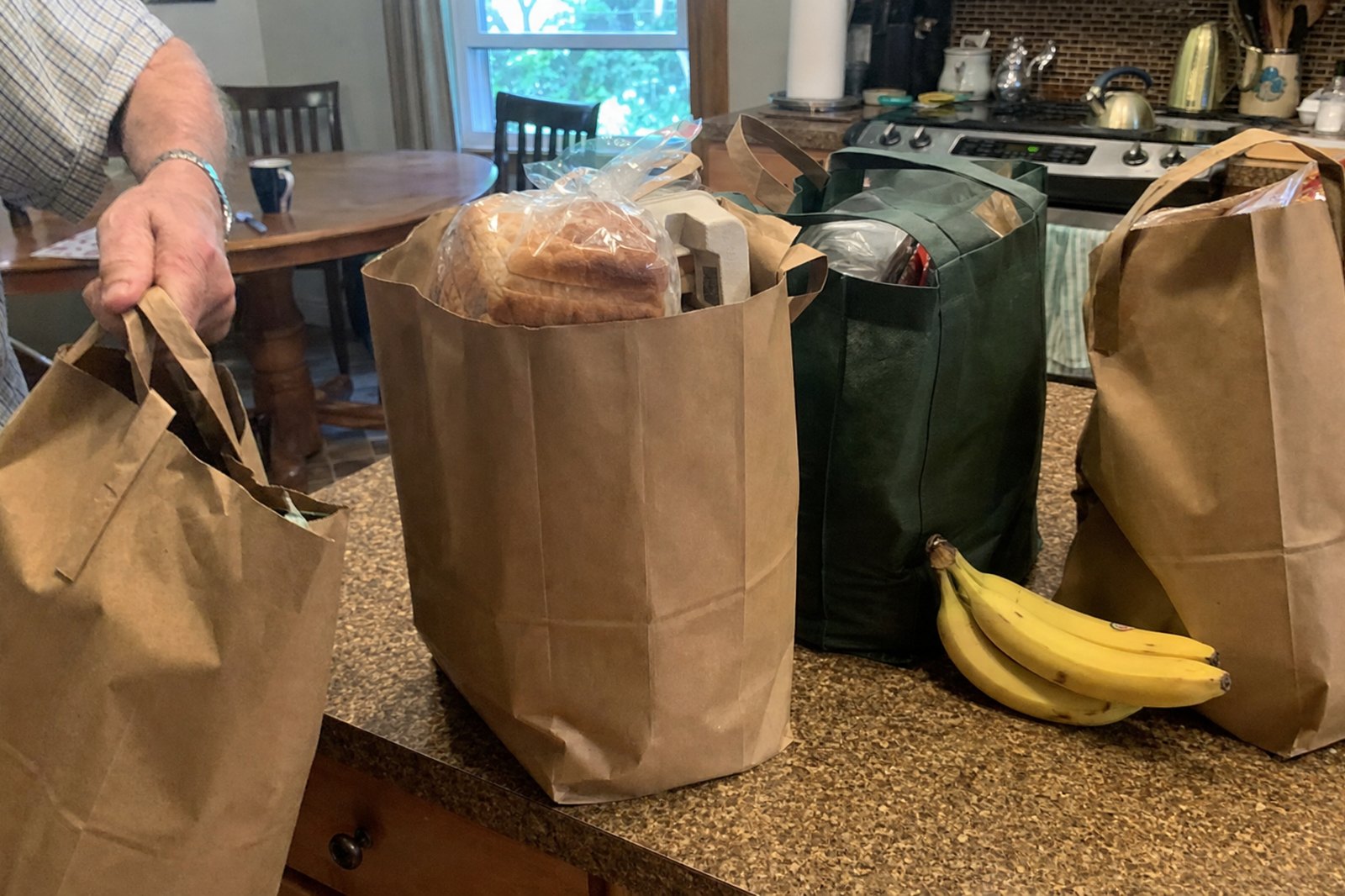 Four grocery bags still full on a kitchen counter, a man's arm setting one more down, the kitchen table out of focus behind.