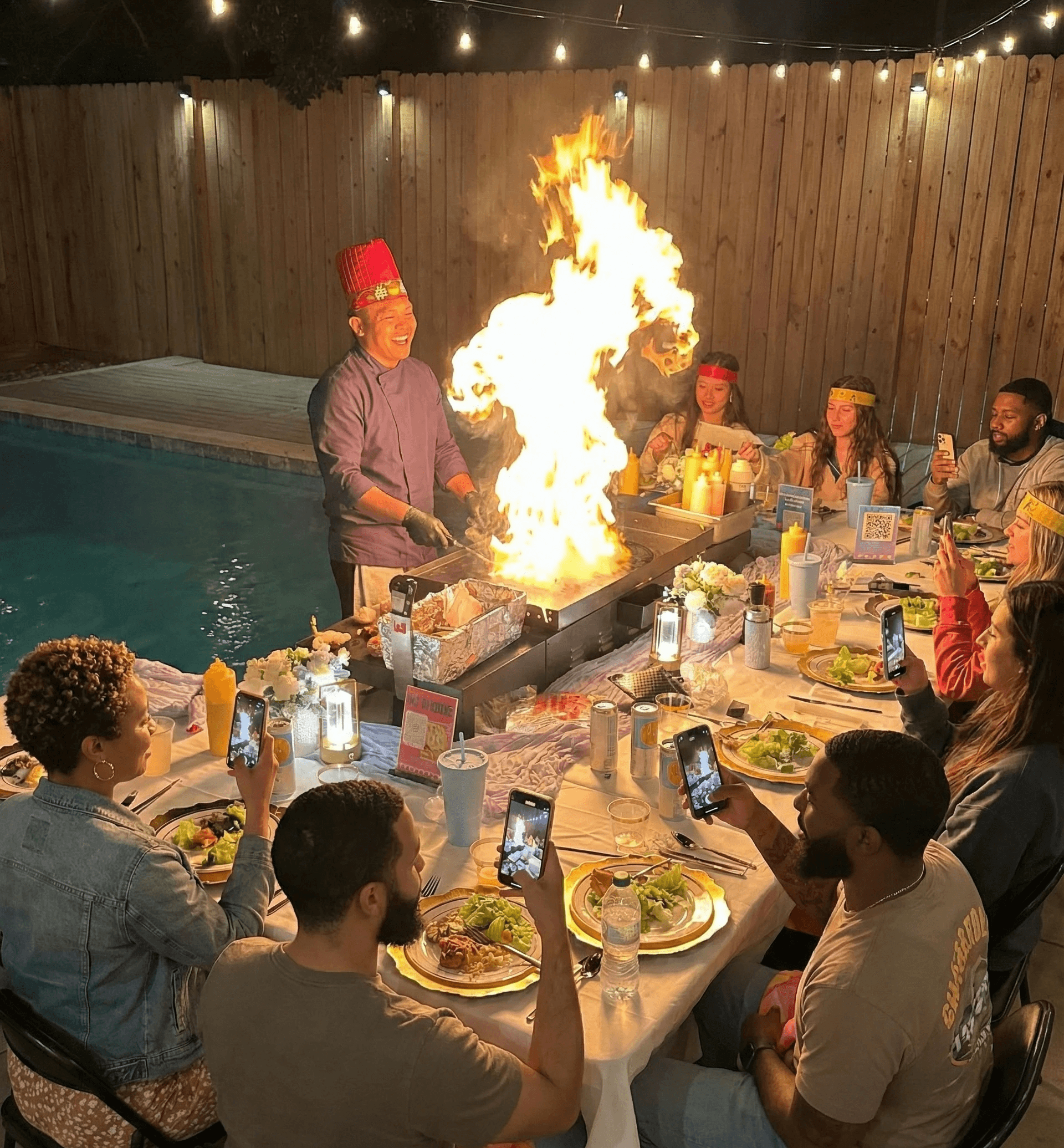 A bright, modern dining room featuring a long light wood table set with several matching wooden chairs and white cushions, illuminated by natural light streaming through large windows overlooking a lush green and sunlit yard.