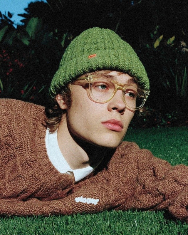 Close-up portrait of a young man with a green beanie and clear-framed glasses lying in the grass, looking directly at the camera.