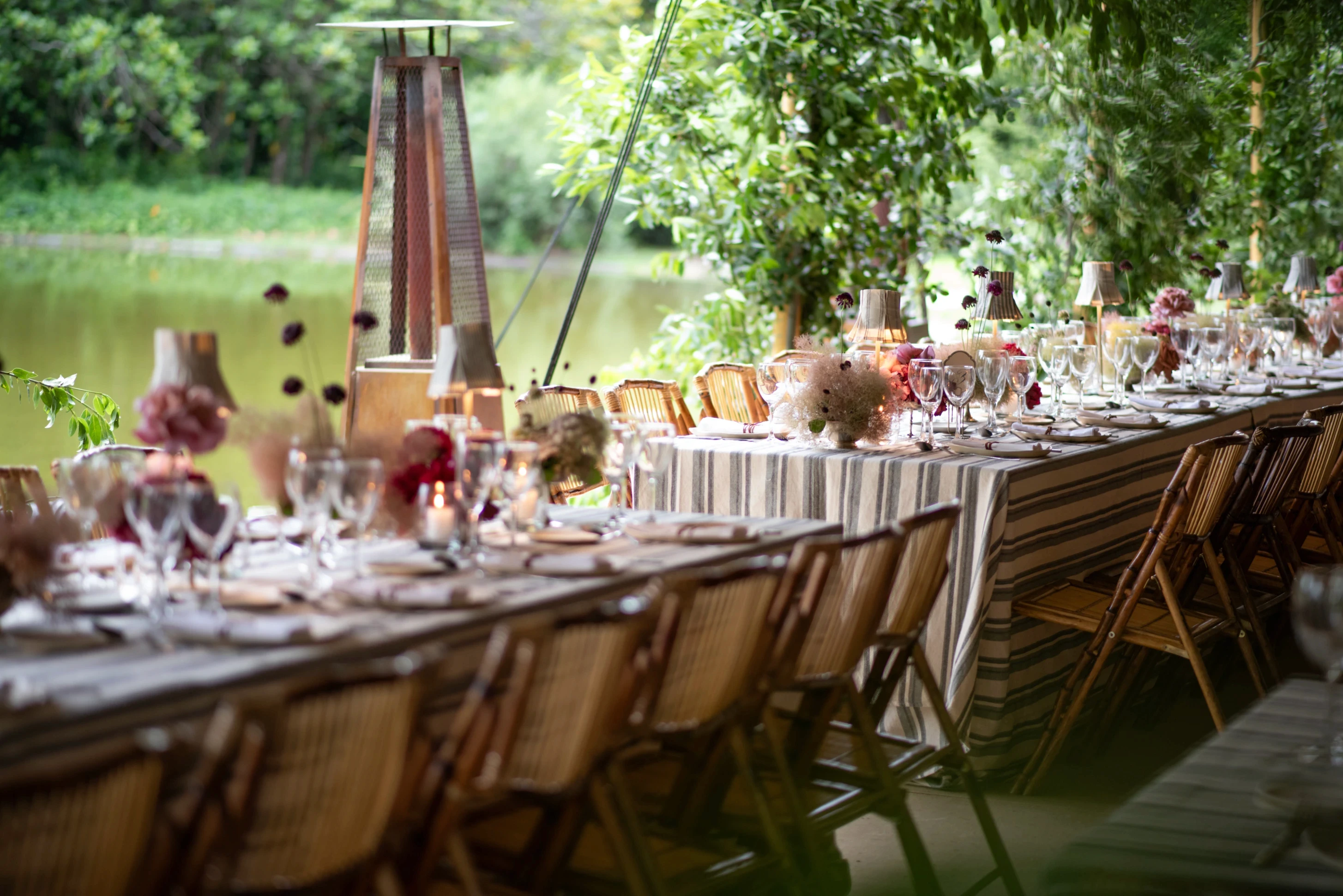 Mesas largas de recepción con manteles a rayas, sillas de caña y lámparas de mesa plisadas en un jardín.