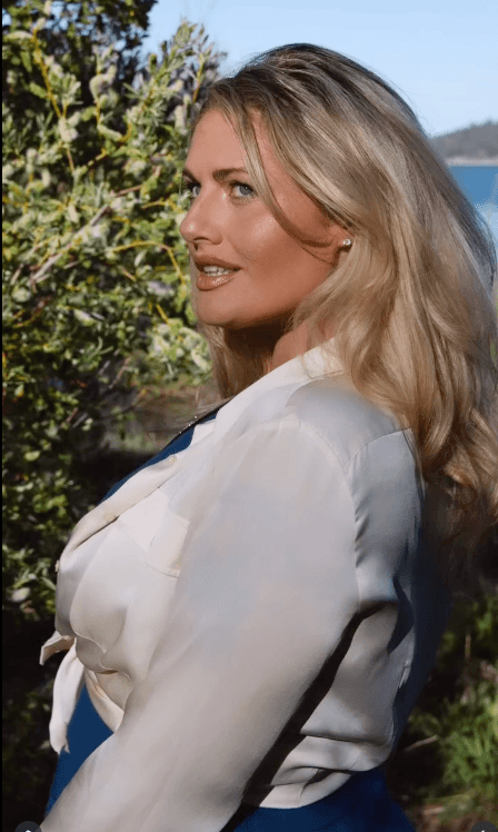 Woman with long blonde hair in nature scenery wearing white and blue outfit.