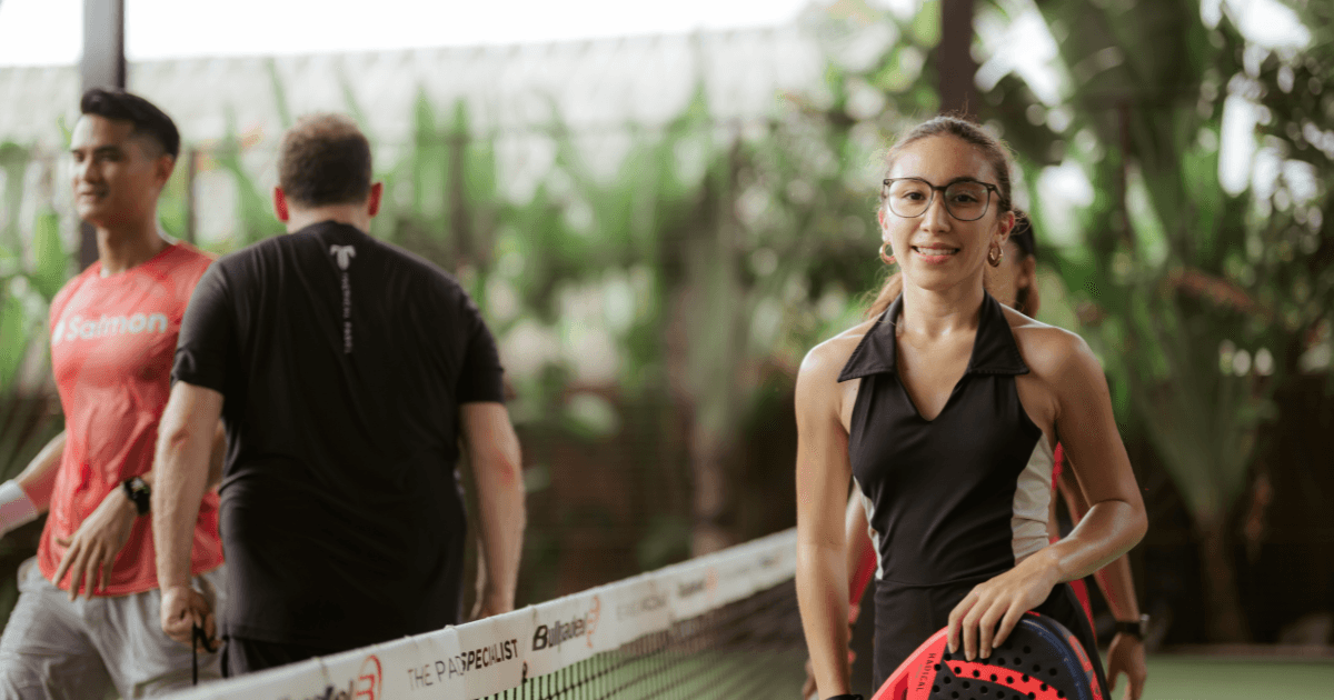 Players smiling and walking off a green padel court surrounded by tropical greenery after a match.