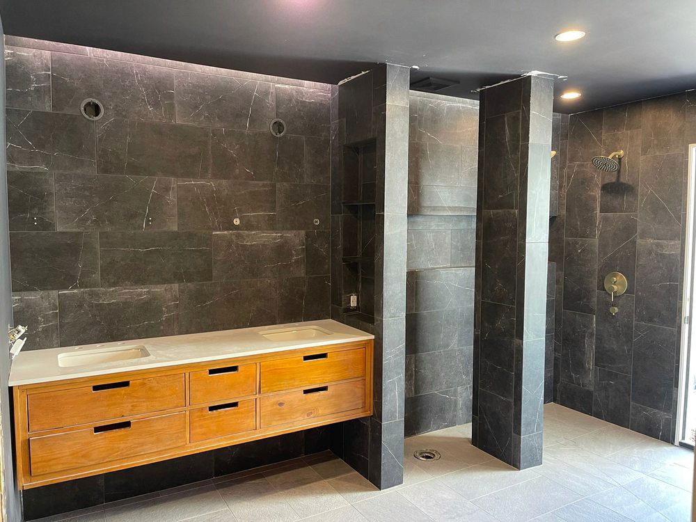 Commercial bathroom tile installation featuring large format dark grey marble-look wall tiles, coordinating tiled columns, and grey floor tiles, alongside a custom wooden vanity.