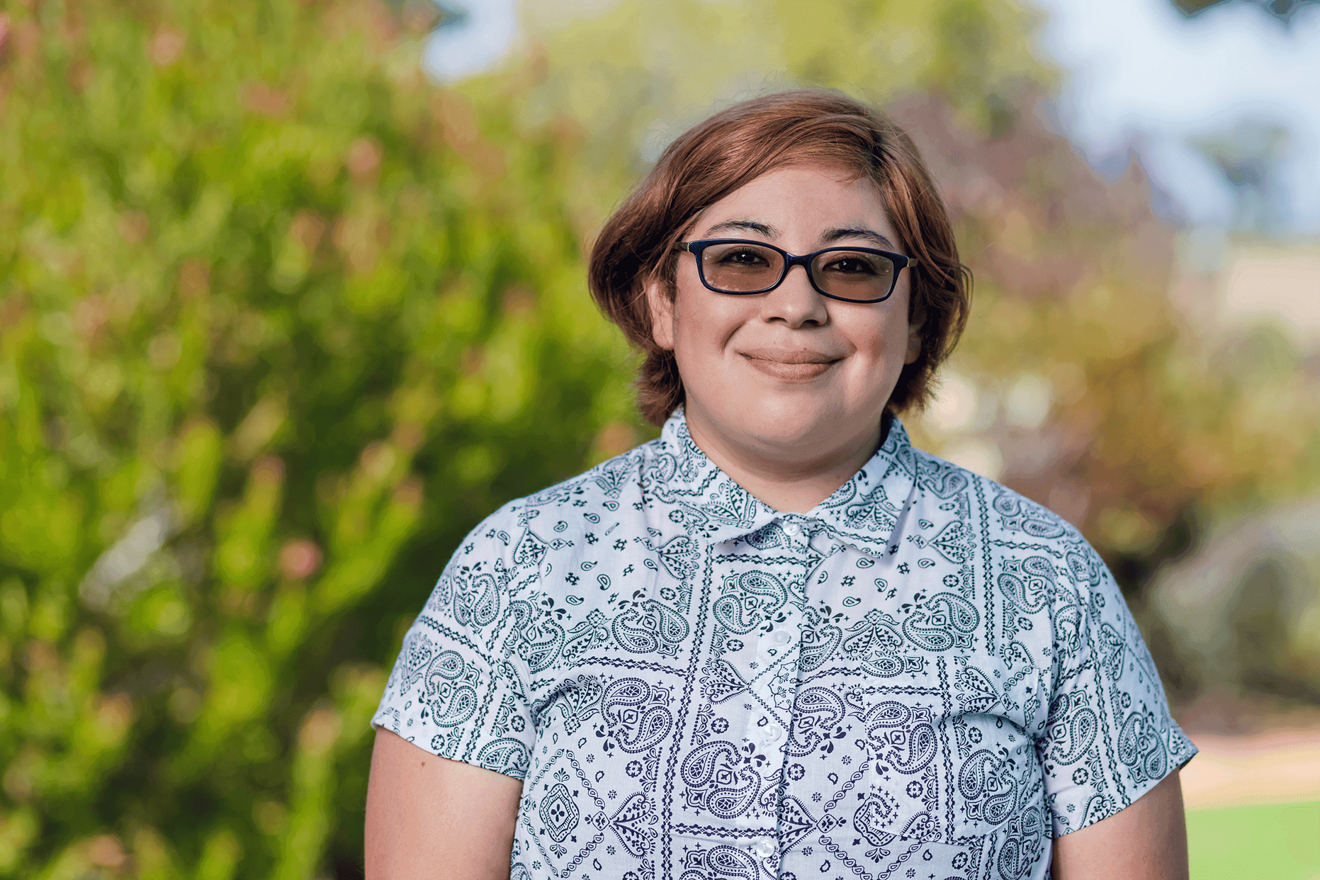 Uma pessoa com cabelo curto castanho-avermelhado e óculos sorri calorosamente, vestindo uma camisa azul estampada, em pé ao ar livre, com um fundo de folhagem verde exuberante.