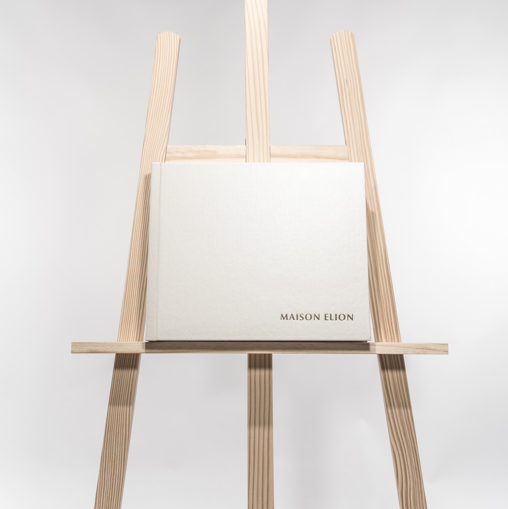 Ivory Fine Art album embossed “MAISON ELION”, poised on a wooden easel — purity elevated to its most silent form. Album Fine Art ivoire embossé “MAISON ELION”, posé sur un chevalet en bois — la pureté portée à son expression la plus silencieuse.
