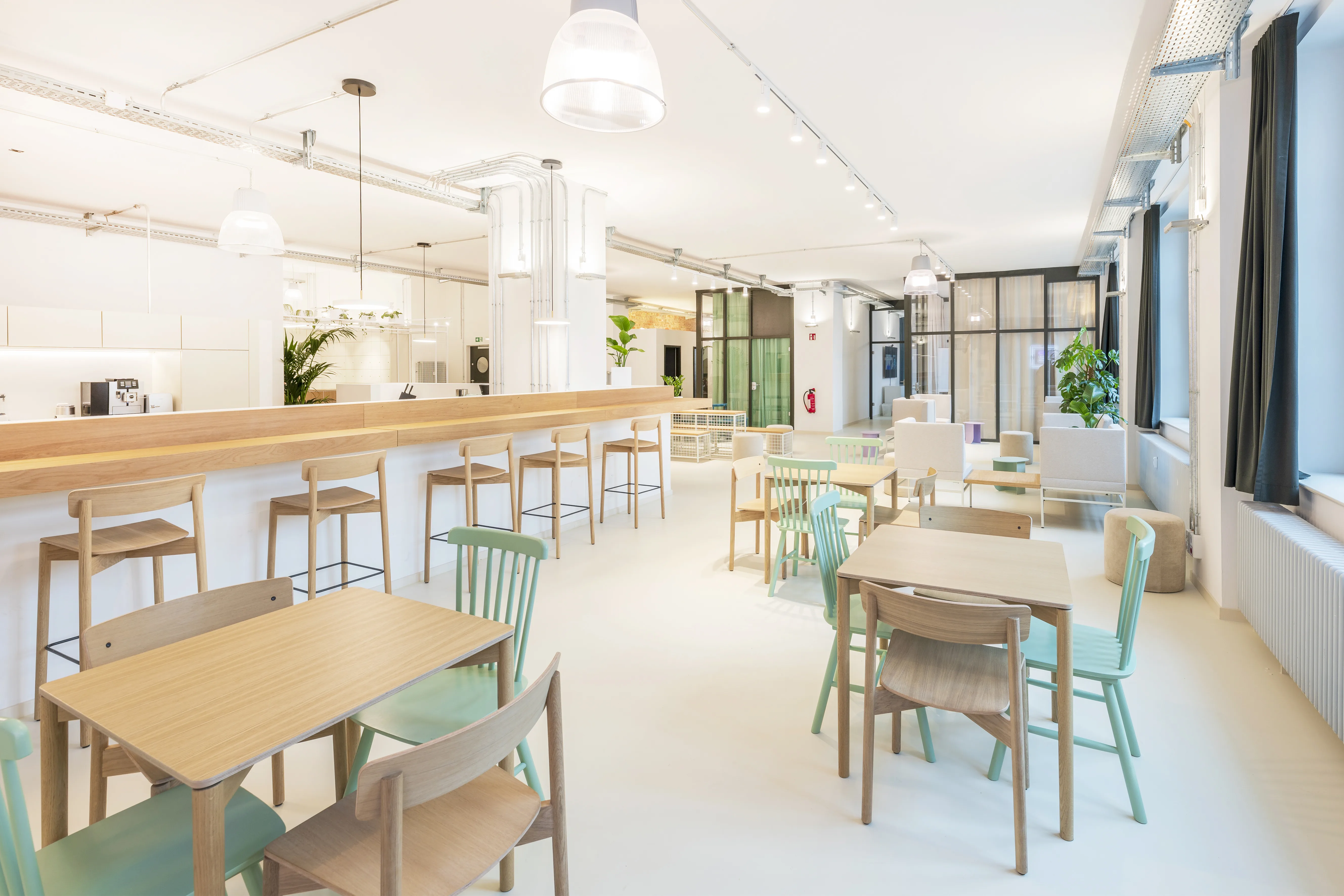 Canteen at ELEMENT with a long oak bar with white stools, wooden tables with mint-green and natural wood chairs, white pendant lights and dark grey curtains along the windows.