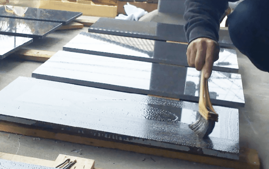 A worker applying a liquid treatment to polished stone slabs using a brush, enhancing the stone's appearance.