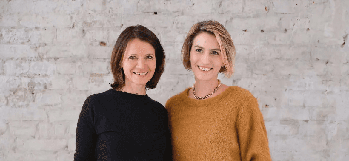 Zwei lächelnde Frauen stehen nebeneinander vor einer hellen Ziegelwand