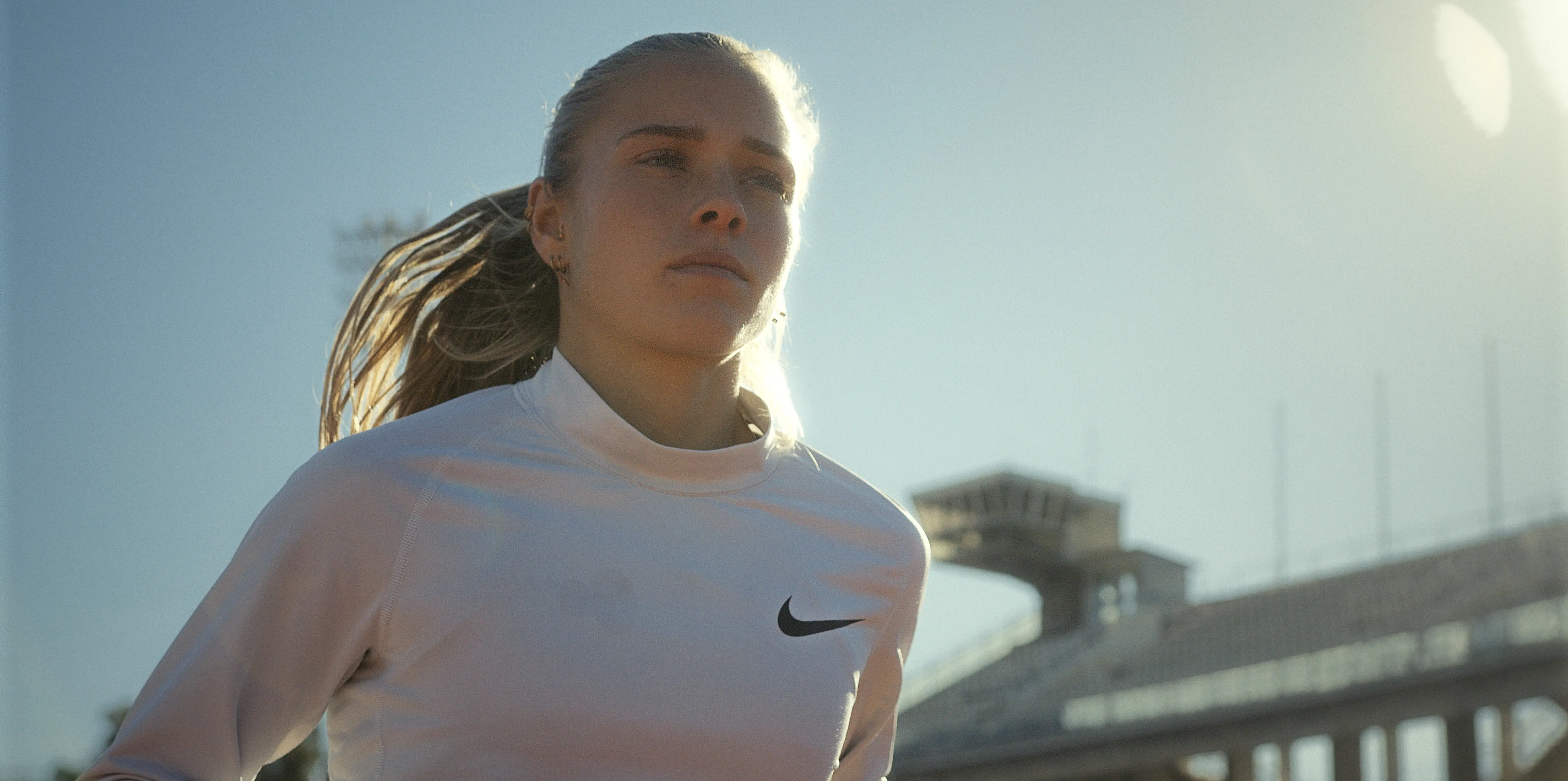 A female athlete wearing a white Nike sweatshirt with her hair tied back is jogging in a stadium during sunrise, casting a warm glow and highlighting the empty bleachers in the background.