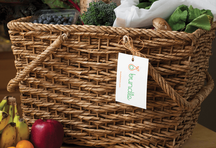 A brown woven wicker basket with two handles sits on a wooden surface. A white tag attached to the basket displays the brand name "Eunado" in green and orange text. Inside the basket are blueberries, broccoli, a white cloth, a brown onion, and a green leaf. A red apple, a yellow banana, and a partially visible orange are placed to the left of the basket.