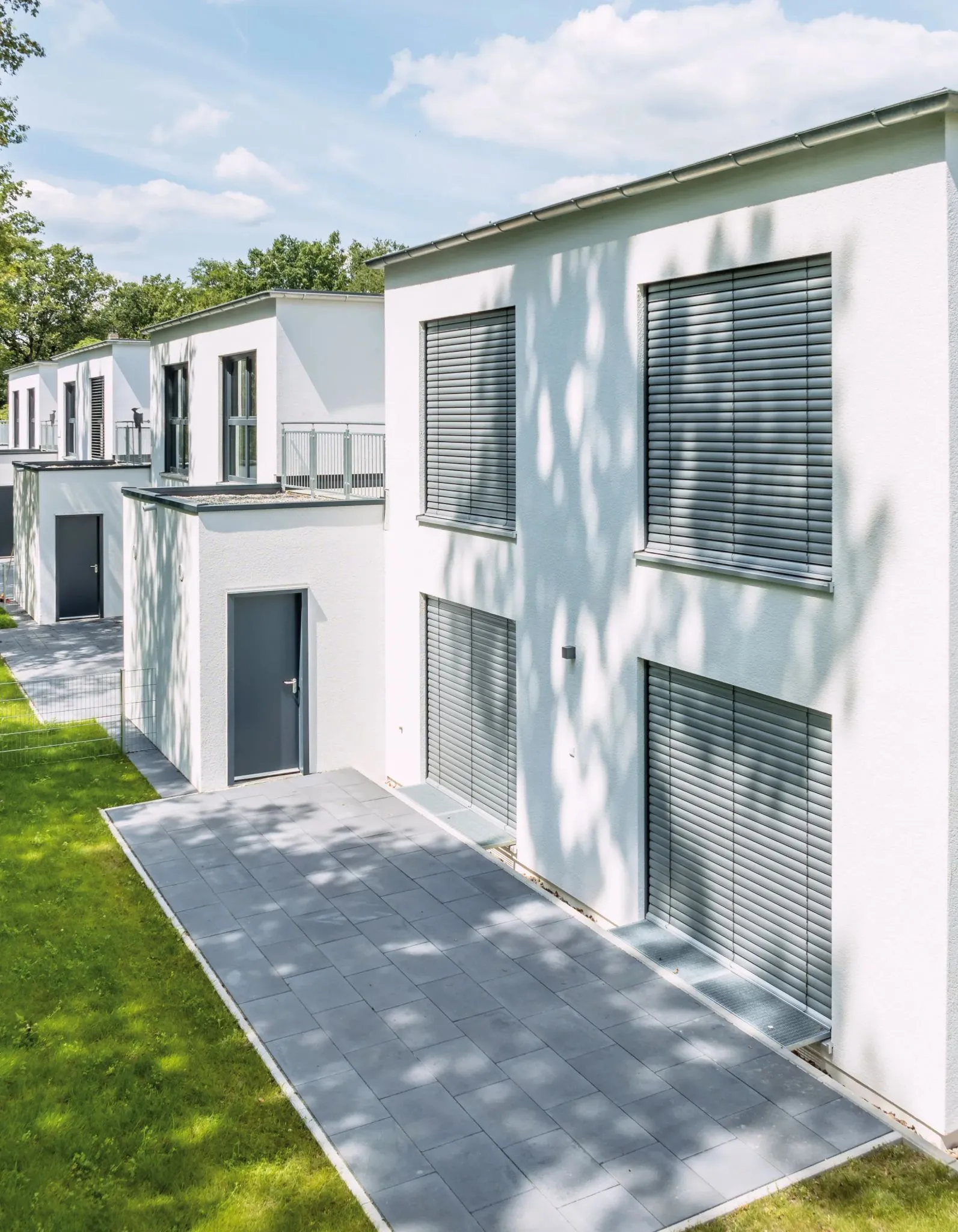 Moderne Reihenhäuser mit weißen Fassaden, grauen Jalousien, grauen Terrassenplatten und grünen Rasenflächen an einem sonnigen Tag.