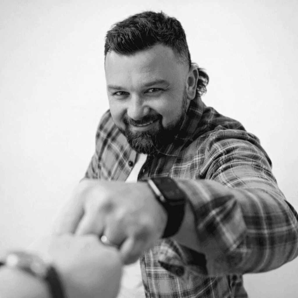 A person with blond hair wears a dark coat and a beige turtleneck sweater. They have a calm expression, against a neutral background.