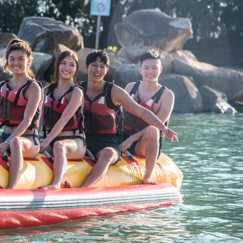 四个身穿救生衣的人坐在湖上的充气香蕉船上，背景是大块岩石和树木。