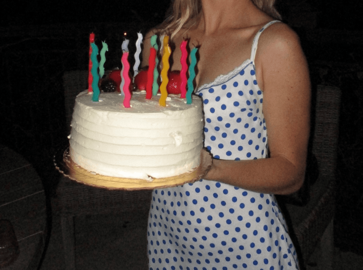 A person holds a birthday cake with colorful candles, wearing a white dress with blue patterns.