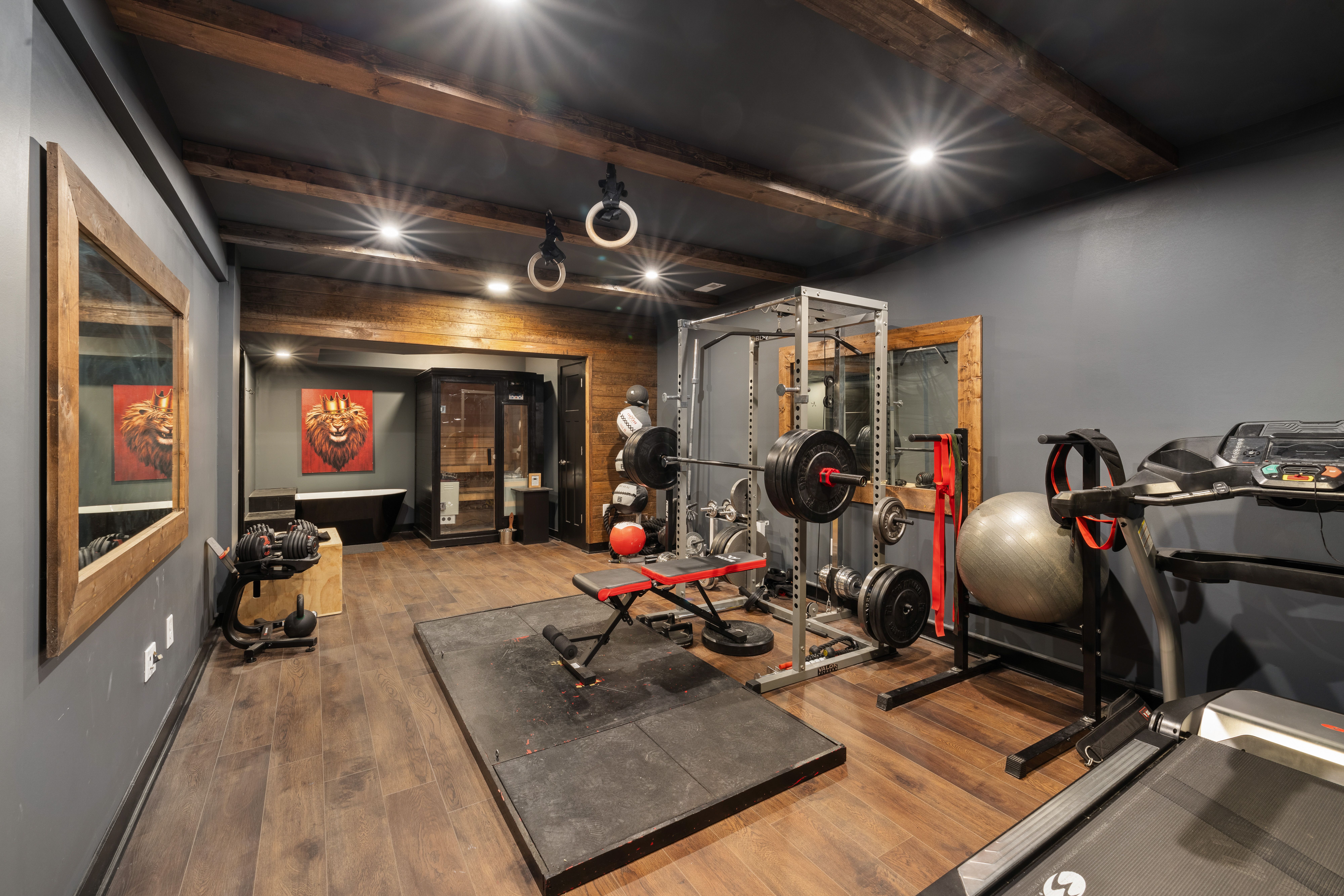 A modern home gym with wooden floors features a variety of fitness equipment, including a red weightlifting bench on a black platform, surrounded by weights, a treadmill, a large exercise ball, and two lion artwork pieces on dark walls.